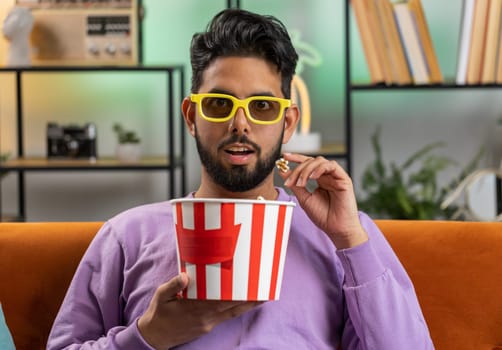 Excited indian man in 3D glasses sits on sofa eating popcorn snacks and watching interesting TV serial sport game, film, online social media movie content at home apartment. Guy enjoying entertainment