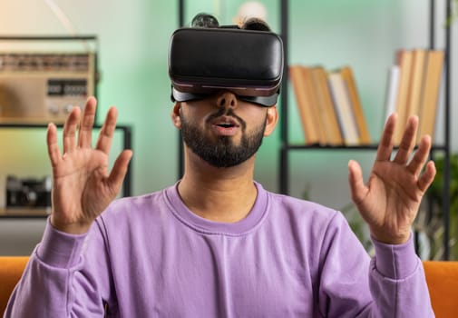 Indian man using virtual reality futuristic technology VR app headset helmet to play simulation 3D 360 video game, watching film movie at modern home apartment. Hindu guy in goggles sitting on couch