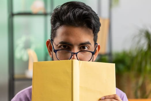 Young indian man in glasses reading interesting book, hiding behind book, enjoying literature, taking a rest on couch. Portrait of peaceful cheerful hindu guy at home apartment living room on sofa