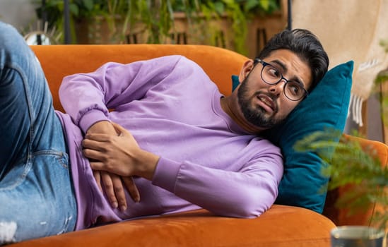 Young indian man lying on sofa feeling sudden strong abdominal stomach ache, gastritis problem. Hindu guy having symptom poisoning diarrhea indigestion peptic ulcer pancreatitis at home apartment room