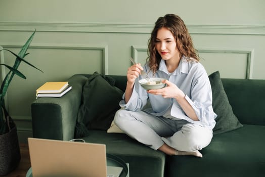 Relaxing weekend at home: Young woman in cozy clothes binge-watching TV series, enjoying leisure time with laptop and snacks on the sofa. A young woman who is taking a break from online learning.