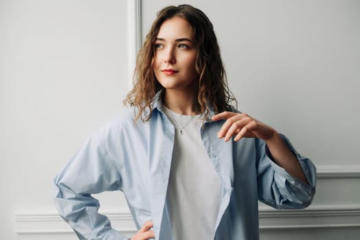 A pensive woman in blue shirt reminiscing about past love, smiling yet with a touch of sadness, standing against a white wall and playing with hair