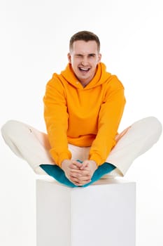young man in orange sweatshirt sitting on white cube on white background. Full length