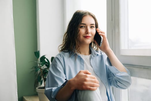 A busy but happy mom enjoying a phone call with her best friend while kids play in the background. Casual indoor lifestyle portrait of a charming 30s woman sitting on windowsill.