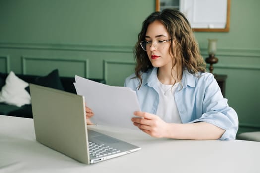 Efficient business woman managing financial data with a laptop and accounting documents in the office. Young female manager or lawyer multitasking with paperwork and laptop at work.