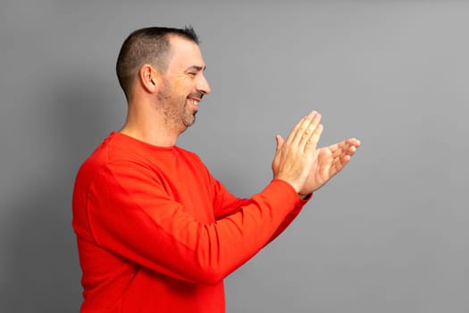 Bearded hispanic man in profile wearing a red jumper feeling happy, smiling and clapping with grateful emphasis for the great show he just witnessed. Isolated on gray studio background.