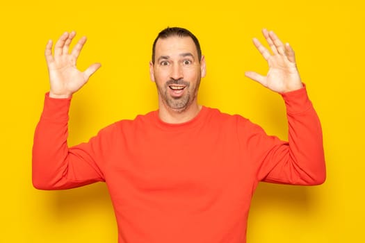 Bearded hispanic man in his 40s wearing a red jumper tremendously surprised by an unexpected visitor, raises his arms excited. Isolated on yellow studio background.