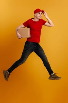 Delivery man in red cap, t-shirt holding cardboard box and jumping isolated on orange background