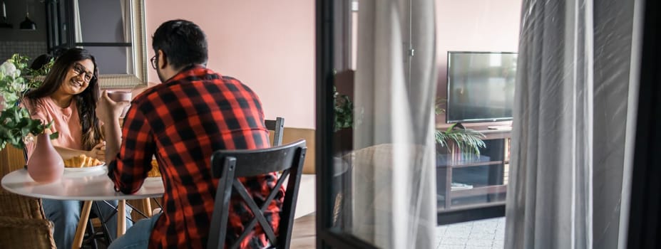 Young diverse loving couple eating croissant and talks together at home in breakfast time. Communication and relationship