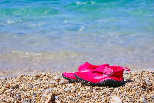 Pair of red swimming shoes on marble pebble beach beside turquoise sea water. Summer holiday concept
