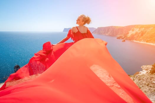 Blonde with long hair on a sunny seashore in a red flowing dress, back view, silk fabric waving in the wind. Against the backdrop of the blue sky and mountains on the seashore