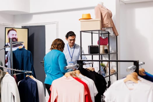 Asian worker showing stylish accessories to woman, discussing items material in clothing store. African american customer shopping for fashionable clothes, checking price in modern boutique