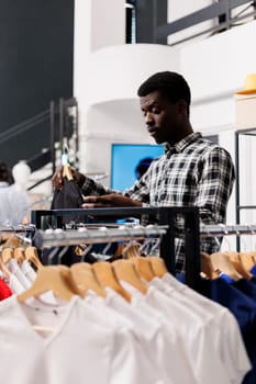 African american customer wanting to buy shirt, looking at colorful clothes in modern boutique. Shopaholic man shopping for casual wear, checking new fashion collection in clothing store