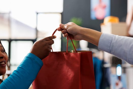 Employee giving red paper bag to customer after purchasing fashionable clothes for new wardrobe in modern boutique. African american woman shopping for casual wear in clothing store