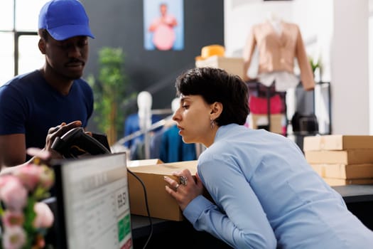 Worker looking at shipping report on tablet computer, giving packages to courier in clothing store. Employee standing at counter desk, preparing customers orders in modern boutique