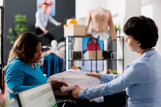 African american woman giving multiple shirts to worker, discussing fabric before buying it in clothing store. Stylish customer shopping for casual wear, purchasing new fashion collection