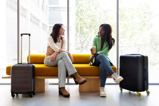 Multiracial young women sitting on hotel reception bench talking. Lifestyle and travel concept.