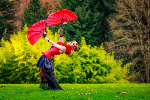 Young beautiful woman in circus costume play with red fans in the park.