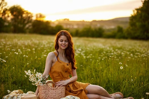 a happy woman in an orange dress sits on a plaid in a chamomile field at sunset and laughs. High quality photo