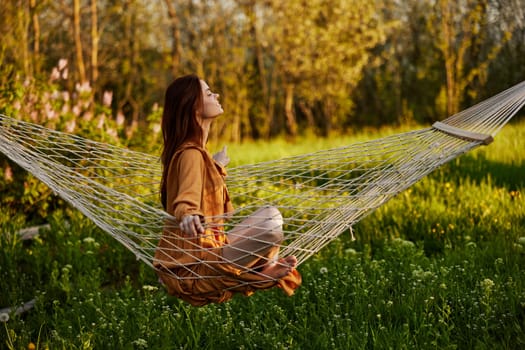 a happy woman in a long orange dress is resting sitting in a hammock at the dacha, smiling pleasantly looking away, illuminated by the summer sun during sunset. Horizontal photo. High quality photo