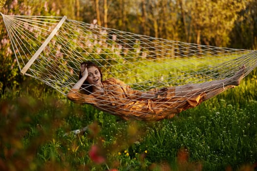 a beautiful woman is resting in nature lying in a mesh hammock in a long orange dress looking at the camera. Horizontal photo on the theme of recreation. High quality photo