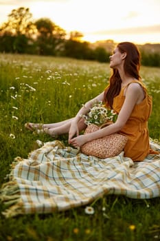 happy redhead woman sits in a field of daisies on a plaid during sunset and enjoys nature. High quality photo