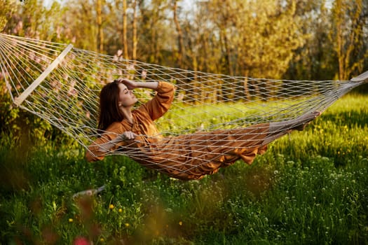 an elegant, slender woman is resting in nature lying in a mesh hammock in a long orange dress enjoying the rays of the setting sun on a warm summer day looking away. High quality photo