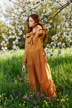 a bright vertical photo of an attractive woman in a long orange dress standing next to a flowering tree in sunny, warm weather, straightening her hair with her hand and closing her eyes. High quality photo