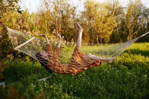 a funny woman is resting in nature lying in a mesh hammock in a long orange dress lifting up her arms and legs. High quality photo