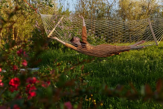 a happy woman in a long orange dress is relaxing in nature lying in a mesh hammock enjoying summer and vacation in the country surrounded by green foliage, happily lifting her legs. High quality photo