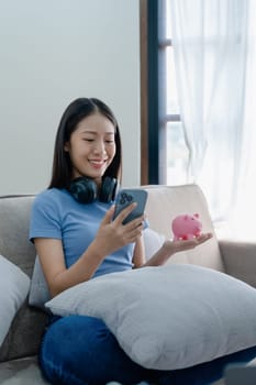 Asian young woman holding a piggy bank and using smartphone mobile, Savings concepts