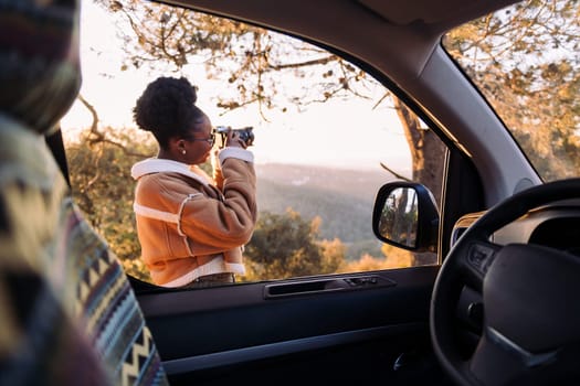 young african woman taking photos of nature with a camera during a road trip in camper van, concept of van life and travel adventures