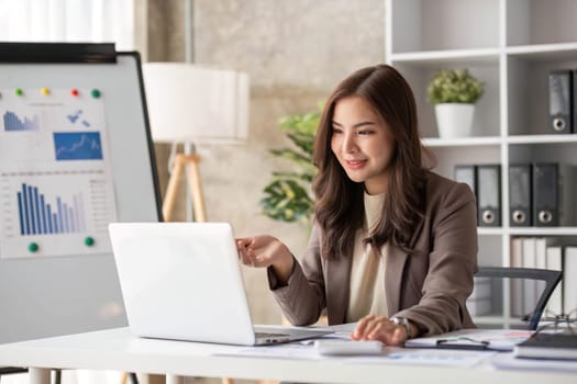 Cheerful business lady working on laptop in office, Asian happy beautiful businesswoman in formal suit work in workplace. Attractive female employee office worker smile...