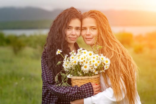 Two women enjoy nature in a field of daisies. Girlfriends hugging hold a bouquet of daisies and look at the camera