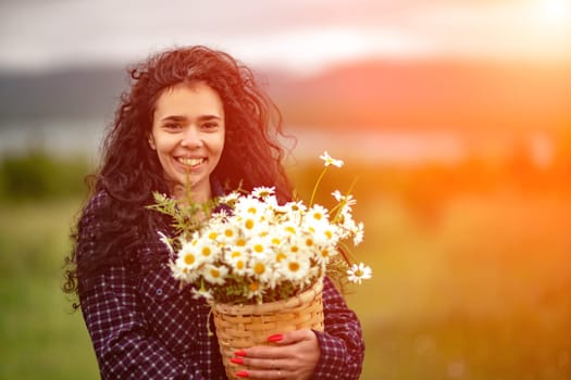 A woman stands on a green field and holds a basket with a large bouquet of daisies in her hands. In the background are mountains and a lake