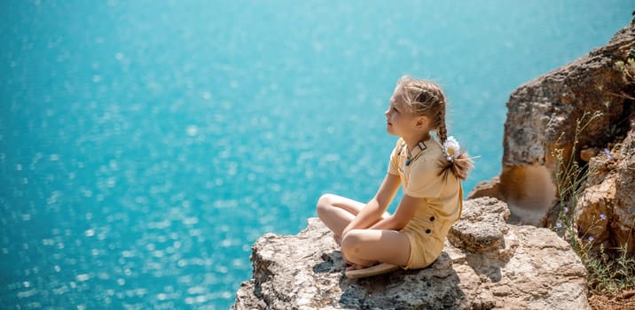 Happy girl perched atop a high rock above the sea, wearing a yellow jumpsuit and braided hair, signifying the concept of summer vacation at the beach