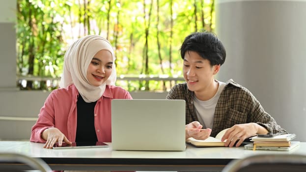 Two friendly university student sitting at table with laptop, talking discussing research, learning tasks together.