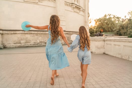 Daughter mother run holding hands. In blue dresses with flowing long hair, they hold balloons in their hands against the backdrop of a sunset and a white building