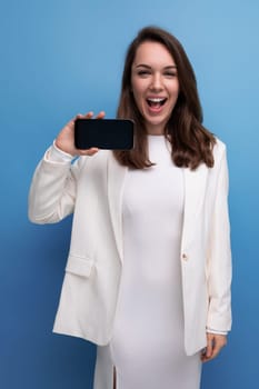 successful energetic young brunette with long hair woman in white dress shows smartphone screen with mockup.
