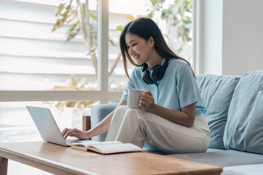 Smiling asian woman in headphones using laptop computer while having her morning coffee sitting on a couch at home.