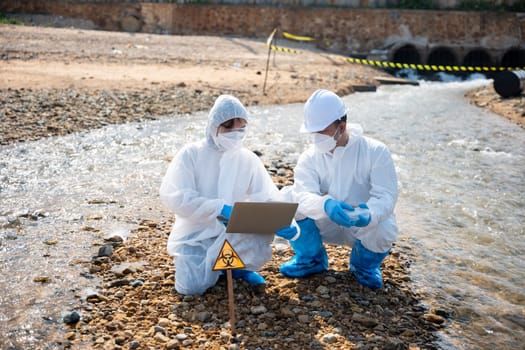 Biologist wear protective suit and mask collects sample of waste water from industry, problem environment, Ecologist sample taken dead fish to inspection and save data to laptop computer, Toxic water