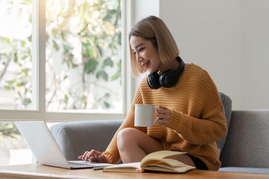 Smiling asian woman in headphones using laptop computer while having her morning coffee sitting on a couch at home.