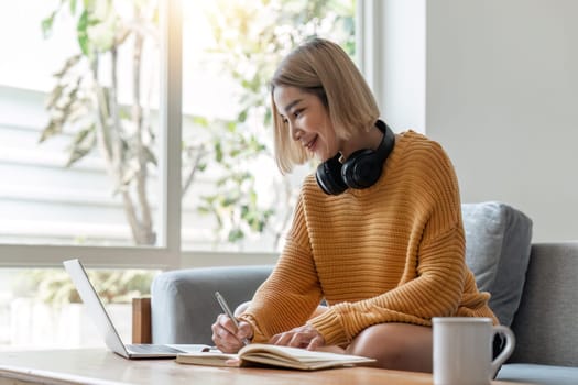 young woman working on laptop and taking notes while listening to lecture at home.