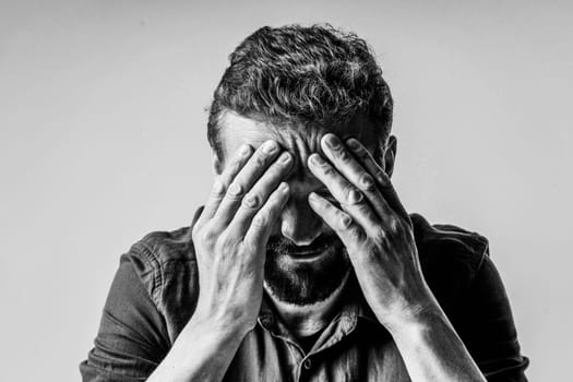 Black and white photo of a man looking extremely sad and depressed. He sitting on white background and has face covered with both hands, showing the despair and hopelessness he feels. High quality photo