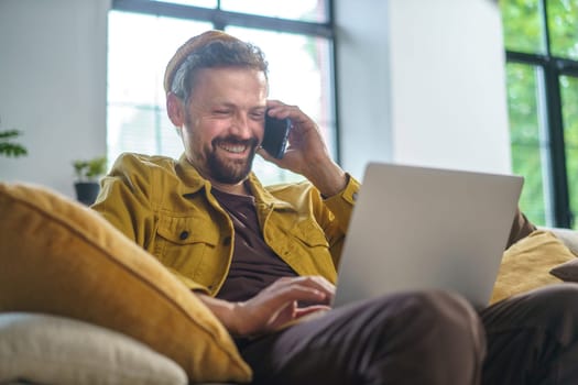Smiling man having conversation on phone while working on laptop on sofa in office. He dressed in casual wear and most likely programmer or IT professional. High quality photo