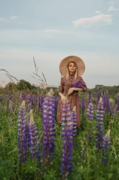 A beautiful woman in a straw hat walks in a field with purple flowers. A walk in nature in the lupin field.