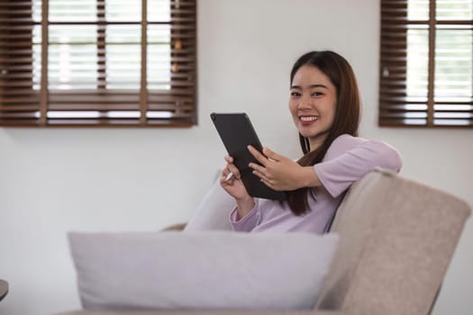 Beautiful attractive young Asian woman sitting on gray sofa and using digital tablet at living room in the morning.