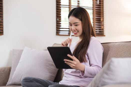 Beautiful attractive young Asian woman sitting on gray sofa and using digital tablet at living room in the morning.