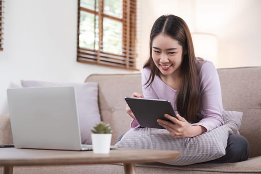 Beautiful attractive young Asian woman sitting on gray sofa and using digital tablet at living room in the morning.