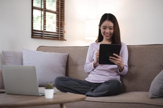 Beautiful attractive young Asian woman sitting on gray sofa and using digital tablet at living room in the morning.